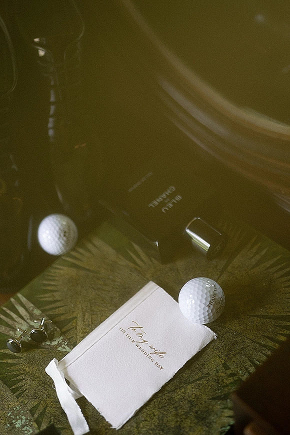 Invitation suite wedding invitation flat lay on a patterned surface, deckle-edge card with cufflinks, perfume, lipstick, and golf balls in warm light
