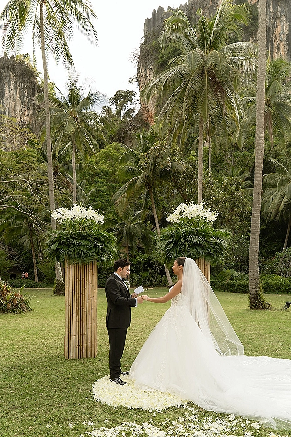 Wedding vows during an outdoor wedding ceremony as bride and groom hold hands beneath white floral altar pillars in a tropical garden setting
