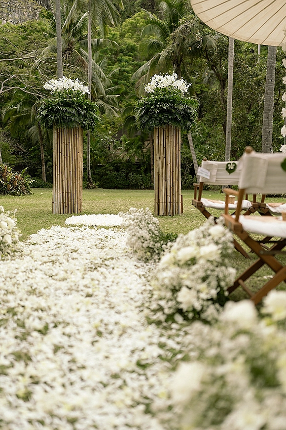 Ceremony aisle decor with a floral aisle runner of white ground arrangements and bamboo pedestals on a grassy lawn with palms in back