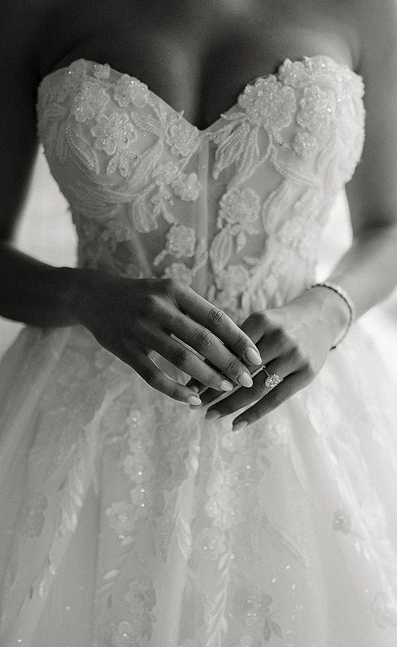 Wedding dress close-up showing strapless lace bodice with floral appliques and beaded embroidery, bride’s hands with engagement ring in soft interior blur