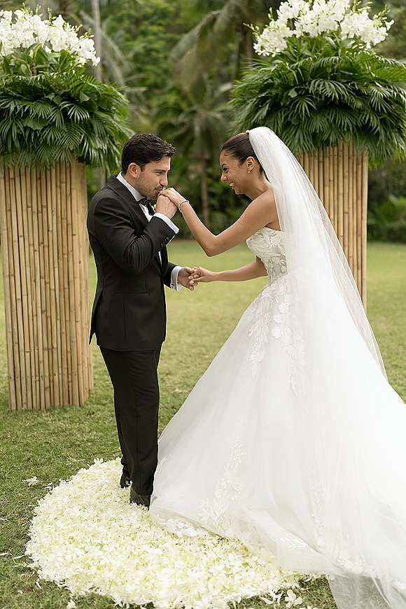 Ceremony moment of groom kissing bride hand as they hold hands on a flower-petal aisle before bamboo panels and palms on a lawn