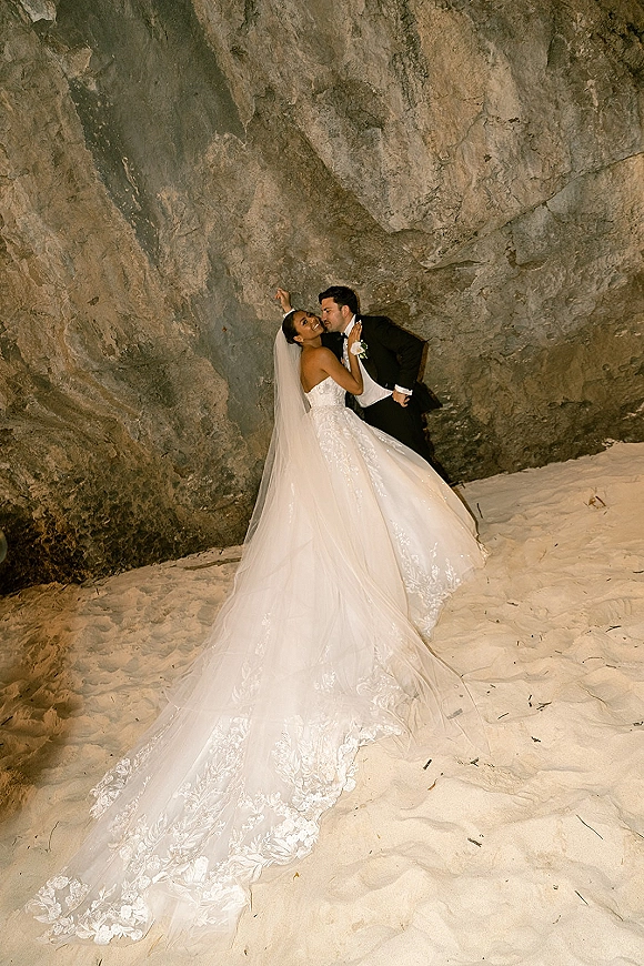 Wedding kiss portrait of the bride and groom kissing, her long veil trailing on sand by a rock cliff, groom in tuxedo with boutonniere
