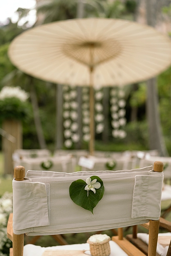 Ceremony seating decor with white canvas director chairs, green leaf chair markers and single white boutonniere in a tropical garden aisle