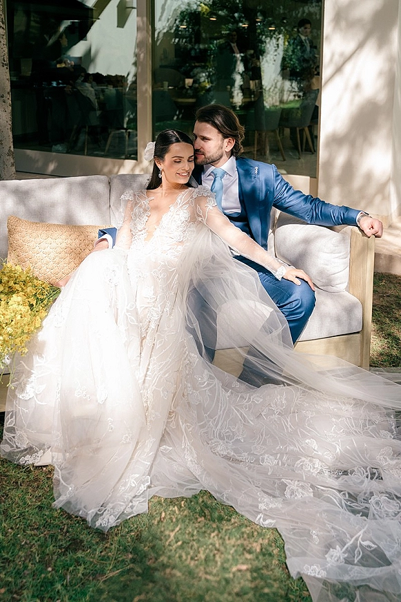Couple portrait of bride and groom on sofa, groom in blue suit kissing her forehead as she wears lace dress and long veil by glass windows
