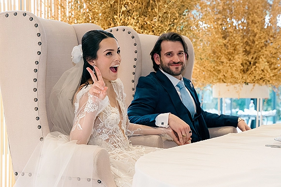 Reception sweetheart table with bride and groom seated, bride in long-sleeve lace gown and veil on wingback chairs before gold foliage backdrop