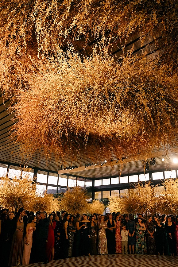 Reception ceiling decor with a hanging floral installation of dried florals and warm string lights above a crowded indoor venue with windows