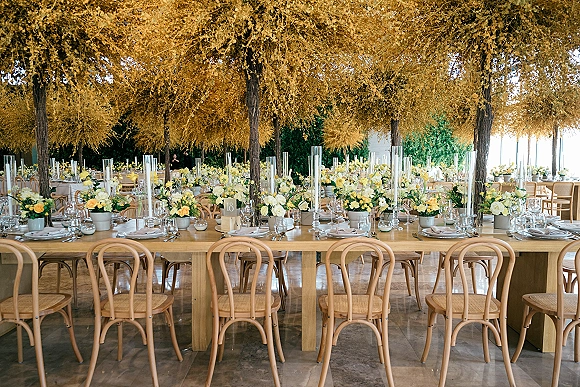 Reception tablescape with long banquet table wedding decor, yellow and white florals, candles, and place settings beneath a floral ceiling installation