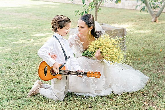 Bride and child portrait with bride holding a yellow wildflower bouquet, veil and lace sleeves beside a ring bearer with guitar on a lawn