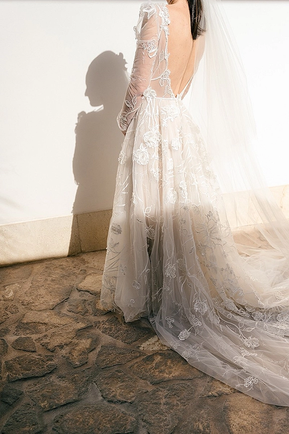 Wedding dress with lace wedding dress details, sheer long sleeves and low back, chapel veil and train in sunlight against a white wall