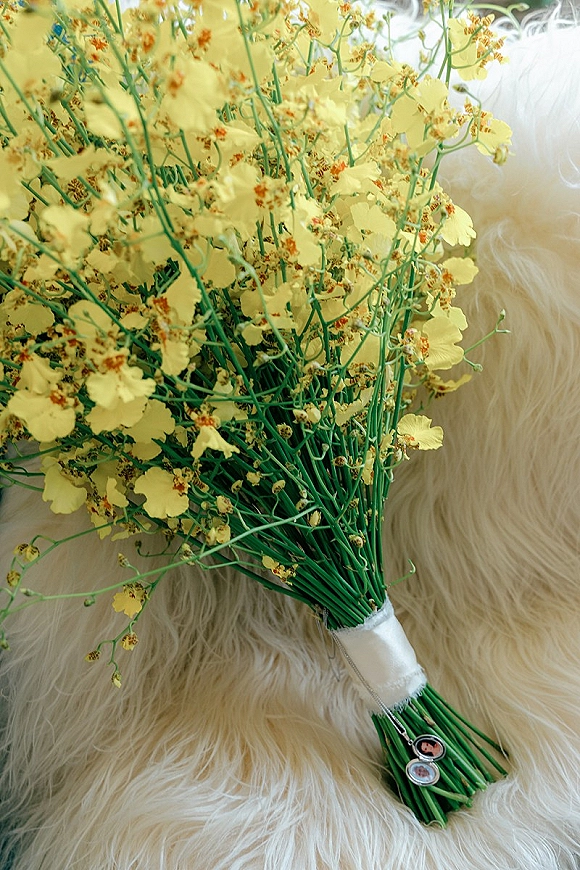 Wedding bouquet with yellow flowers and green stems, wrapped in white ribbon with pins, resting on soft white faux fur background