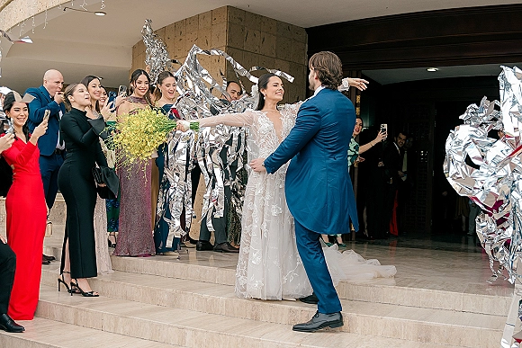 Wedding exit with newlyweds stepping down marble stairs as guests wave silver foil streamers; bride in lace gown and veil holds bouquet