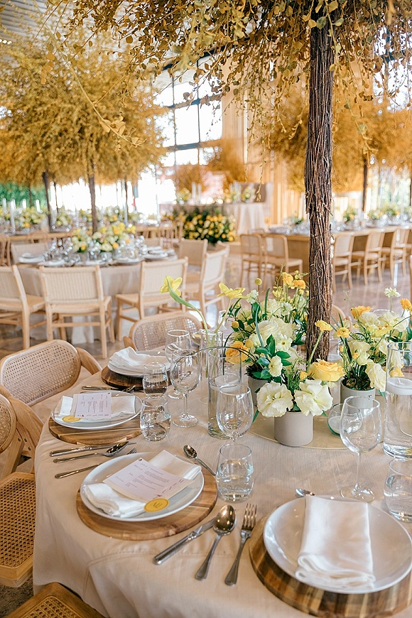 Reception tablescape with round wedding table setup featuring wood charger plates, white napkins, yellow blooms, and candles under hanging greenery in a glasshouse