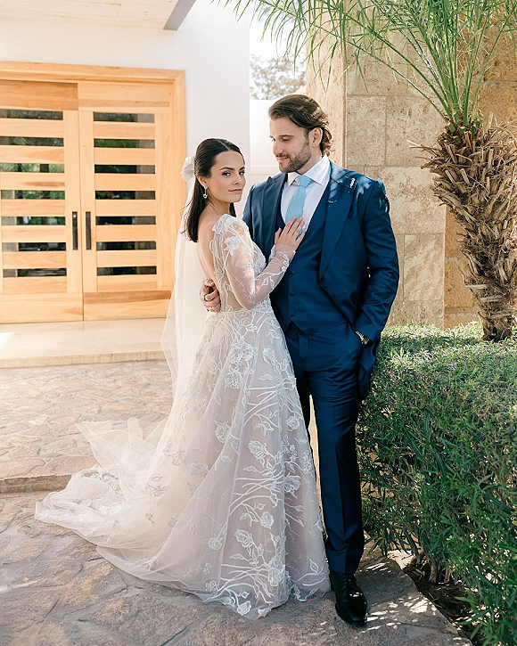 Couple portrait of bride in long-sleeve lace gown and veil embracing groom in a navy suit by stone wall and wooden doors outdoors