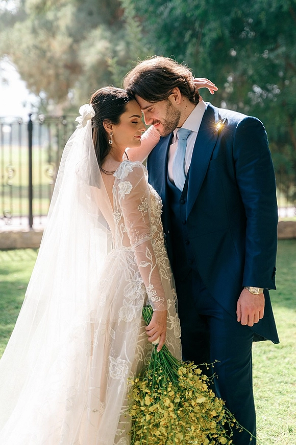 Couple portrait of bride and groom embrace with forehead touch, her long veil and wildflower bouquet in an outdoor garden by fence