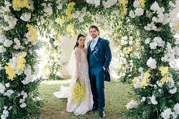 Couple portrait of bride in lace dress and veil with cascading bouquet beside groom in blue suit under a white and yellow floral arch on a garden lawn