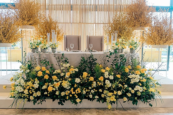 Sweetheart table decor with yellow roses and white flowers, candles in glass cylinders, and tall dried florals against a wood slat backdrop
