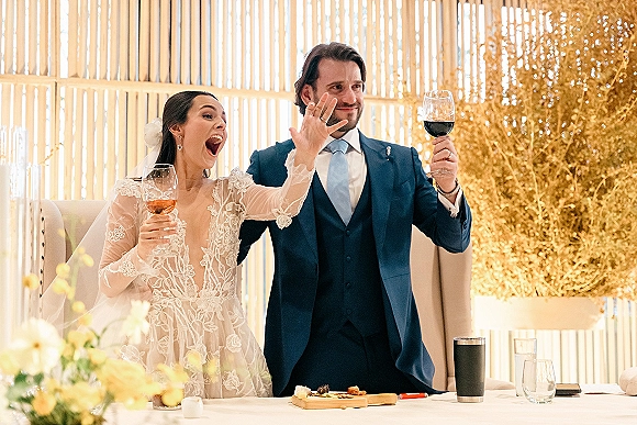 Wedding toast as bride and groom raise red wine and rosé glasses at their sweetheart table, lace sleeves and veil before dried florals