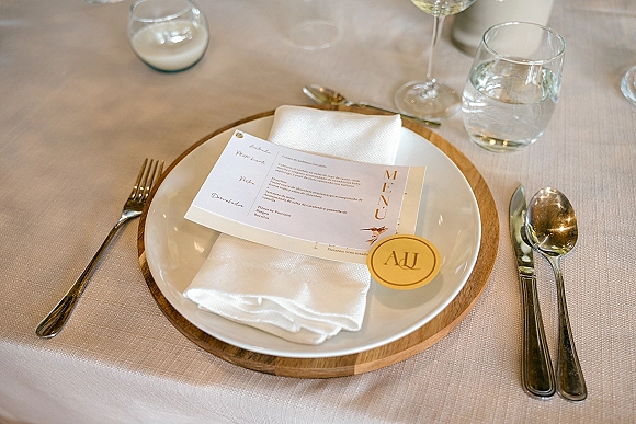Wedding place setting with wedding menu card on a white napkin and plate atop a wooden charger, with gold wax seal, glasses, and candle on linen cloth