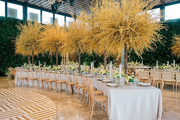 Reception tablescape with a long banquet table wedding setup, white linens, tall yellow floral centerpieces and taper candles by a green hedge wall