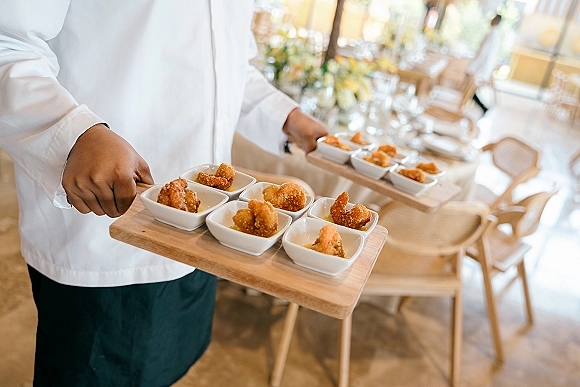 Wedding passed appetizers on wooden trays held by a server in uniform, with white tasting bowls at indoor reception tables by sunny windows