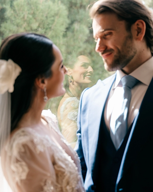 Couple portrait of bride and groom portrait gazing at each other in window light, her lace sleeves and veil softly backlit with greenery behind