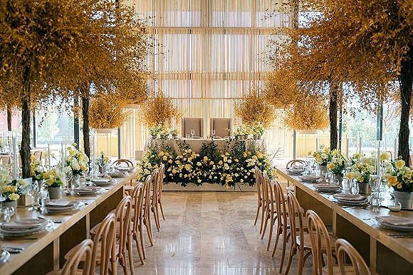 Reception tablescape with long banquet tables set with glassware, candles, and yellow and white florals under a dried foliage installation by tall windows