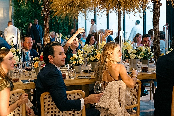 Wedding reception toast with wedding guests cheering, raised glasses along a wood banquet table with candles, florals, and evening windows