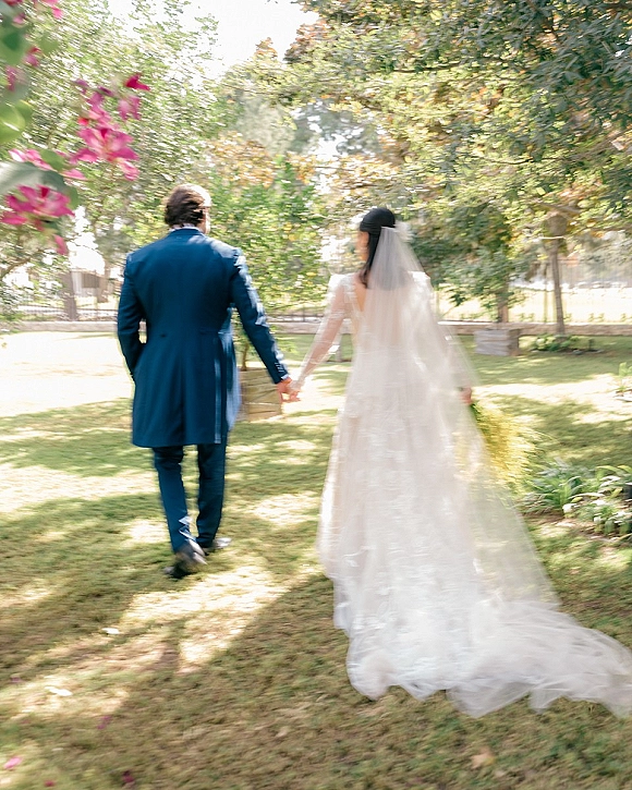 Wedding couple walking away hand in hand, bride and groom walking away with a long veil trailing across a sunlit garden lawn by trees