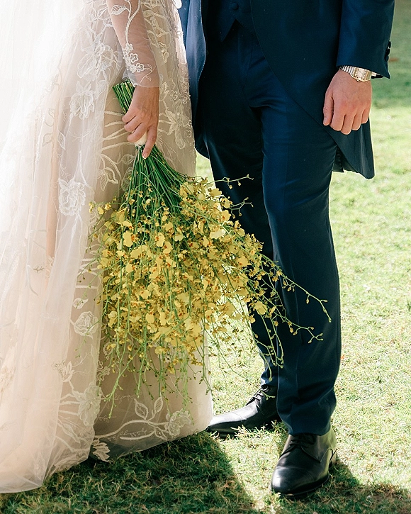 Wedding couple portrait with bride holding cascading wildflower bouquet, lace sleeves and long veil beside groom in suit on grass lawn