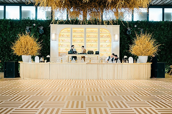 Wedding bar setup with a curved counter, backlit shelves, glassware and cocktail bottles, framed by a hedge wall backdrop and uplighting