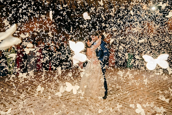 First dance as bride and groom dancing on the reception dance floor under string lights, wedding guests watching amid butterfly confetti