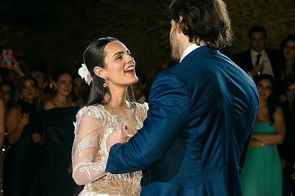 First dance as bride in a lace long-sleeve gown laughs with groom in a blue suit on a dimly lit reception floor as guests watch