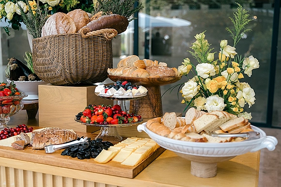 Wedding brunch buffet with bread baskets, cheese slices, berries and dried fruit on wooden risers, with greenery florals by glass windows