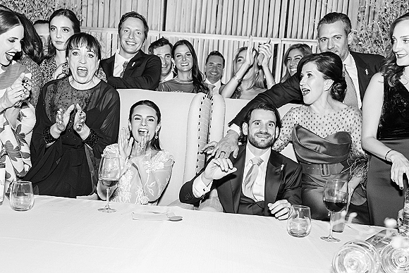 Wedding reception guests in formal dresses and suits laugh and clap on an upholstered sofa, wine glasses on tables by a wood slat wall.