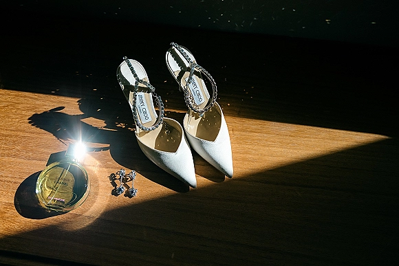 Bridal shoes in a wedding detail flat lay, white pointed-toe heels with rhinestone ankle straps beside perfume and drop earrings on sunlit wood floor