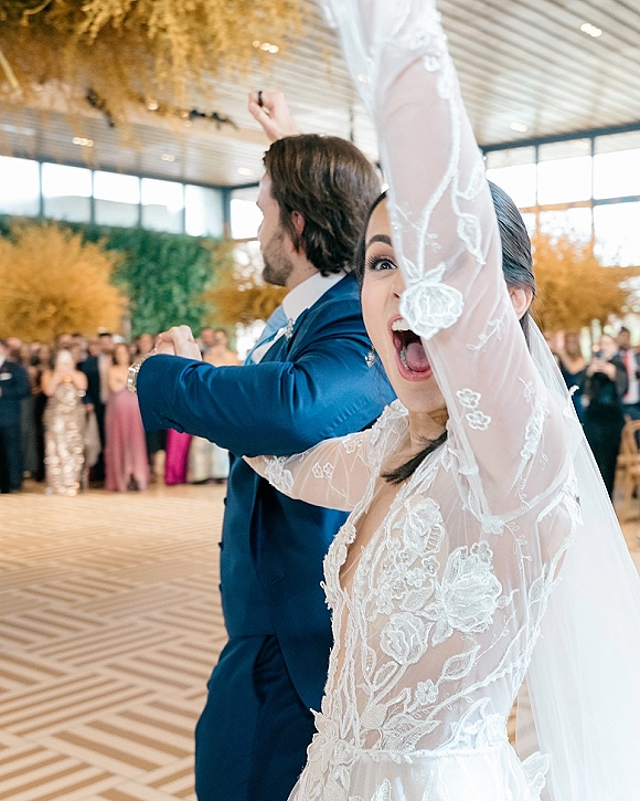 Wedding reception entrance as bride in long-sleeve lace dress and veil raises her arms beside groom in blue suit on ballroom dance floor