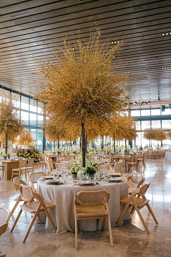 Reception tablescape with a tall tree centerpiece, yellow floral branches, and bud vases on a round white-linen table in a modern hall with large windows