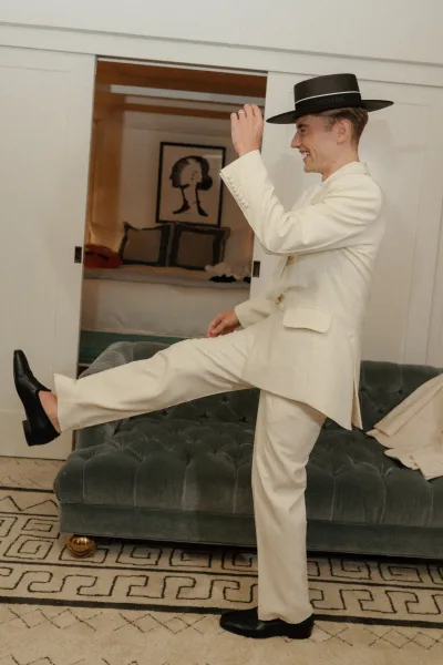 Groom portrait in a white suit with a black hat and black loafers, standing in a bedroom with bed, wall art, and sliding doors