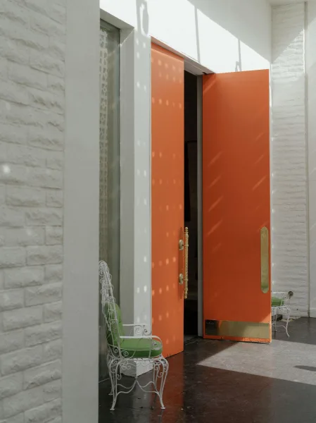 Wedding venue entrance with orange wedding doors and brass hardware, paired with a wrought iron chair and green cushion by white brick walls