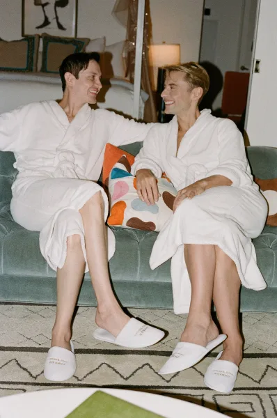 Couple portrait of two grooms in getting ready robes and spa slippers laughing on a sofa in a hotel suite living room