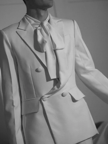 Bridal suit in a white bridal suit with bow necktie, tailored blazer and buttons, posed against a plain wall with soft shadows