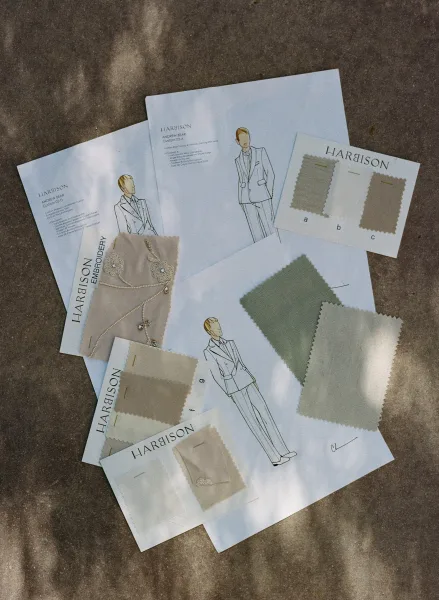 Suit fabric swatches laid out with suit sketch illustrations on paper stationery in dappled sunlight across a concrete ground