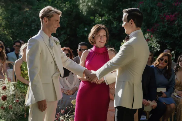 Ceremony moment at an outdoor wedding ceremony as two grooms hold hands before an officiant in a bright dress, guests in garden chairs behind