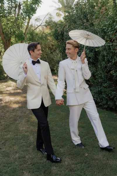 Couple portrait of same sex wedding couple holding hands with a white parasol, walking on a sunny garden lawn with trees behind them