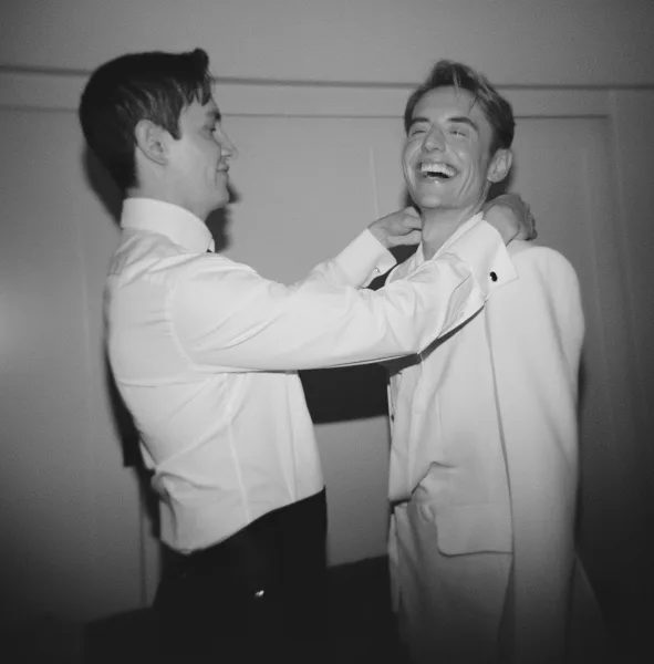 Groomsmen moment as a groomsman adjusts the groom getting ready tie over suspenders and dress shirt against a simple indoor wall