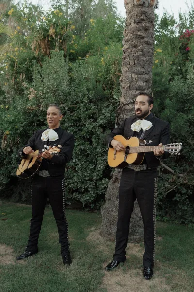 Wedding musicians in mariachi wedding band suits play guitars on a lawn with palm tree and garden shrubs behind them