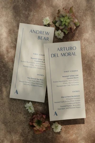 Wedding menu cards with blue ink typography arranged on a stone surface with succulent and floral petal accents in a flat lay