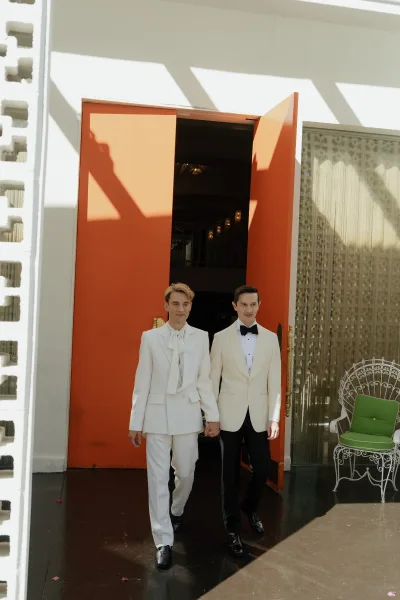 Couple portrait of same sex wedding couple holding hands in white suits by an open door with brass handles, sunlight and shadows on wall