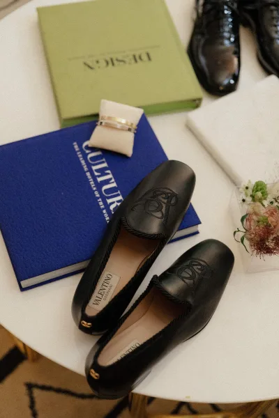 Wedding shoe flatlay with bridal shoe flatlay black leather flats, wedding rings in a ring box, perfume, invitation card, books and flowers on a white tabletop