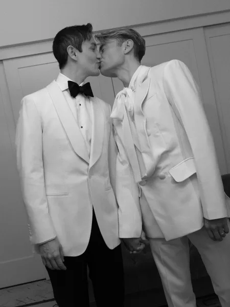 Wedding kiss between two grooms kissing in matching white tuxedos with black bow ties, holding hands indoors by a door and sofa